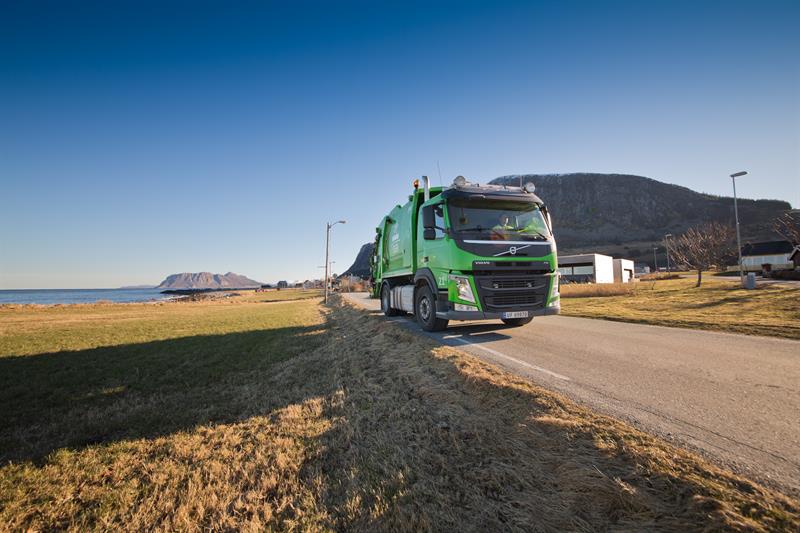 Renovasjonsbil på Floe Foto av ein grøn renovasjonsbil som køyrer på vegen. Den er vinkla delvis mot oss, som at den vil passere oss på høgre side.